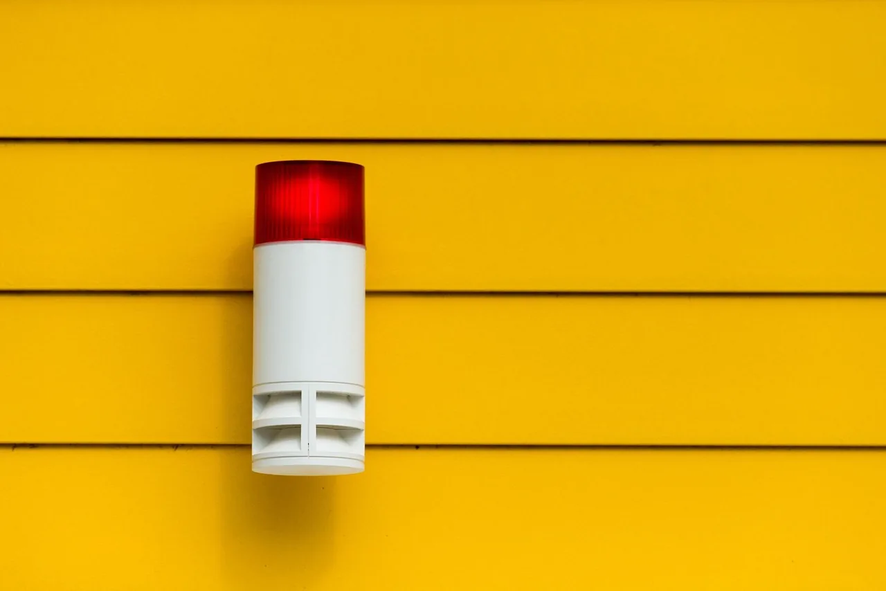Wall-mounted security alarm sounder with a red warning beacon on a yellow exterior wall